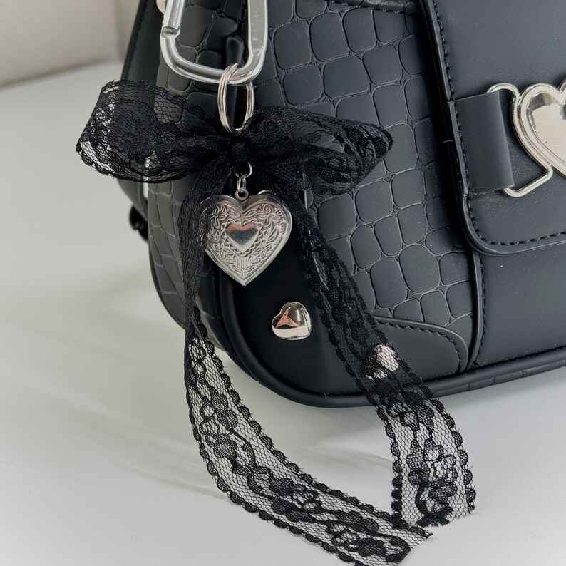 Peut inclure: Sac &agrave; main noir en similicuir avec une texture crocodile. Le sac est orn&eacute; d'un fermoir argent&eacute; en forme de c&oelig;ur et d'un n&oelig;ud d&eacute;coratif en dentelle noire avec un pendentif en forme de c&oelig;ur. Un mousqueton argent&eacute; est attach&eacute; en haut.