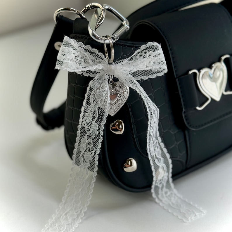Peut inclure: Sac &agrave; main noir avec une texture crocodile, orn&eacute; de clous argent&eacute;s en forme de c&oelig;ur et d'un n&oelig;ud en dentelle blanche. Un charm en forme de c&oelig;ur argent&eacute; est suspendu au n&oelig;ud, attach&eacute; &agrave; un mousqueton argent&eacute;.