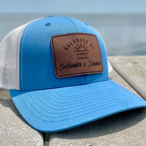 May include: A blue and white trucker hat with a brown leather patch that says "Balanced by Saltwater & Sunshine".