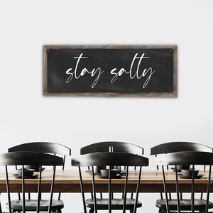 May include: A rectangular chalkboard sign with the words "stay salty" in white script. The sign has a dark gray background and a wooden frame. The sign is hanging above a wooden table and black chairs.