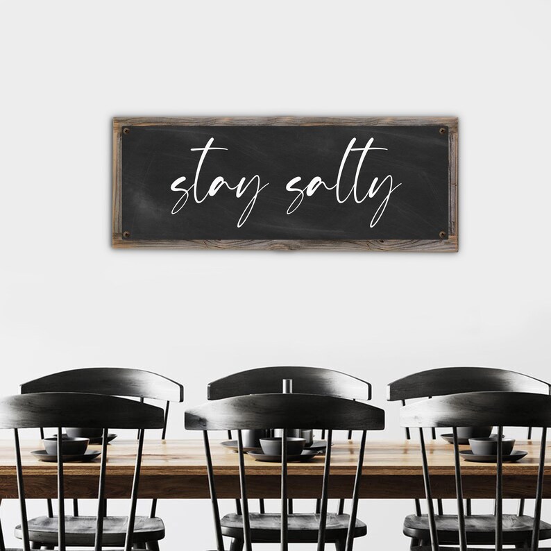 May include: A rectangular chalkboard sign with the words "stay salty" in white script. The sign has a dark gray background and a wooden frame. The sign is hanging above a wooden table and black chairs.
