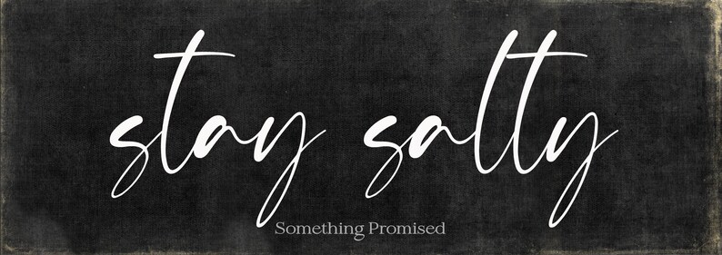 May include: A rectangular black sign with the words "stay salty" in white cursive script. Below the phrase, the text "Something Promised" is written in a smaller, sans-serif font. The sign has a distressed, textured appearance.