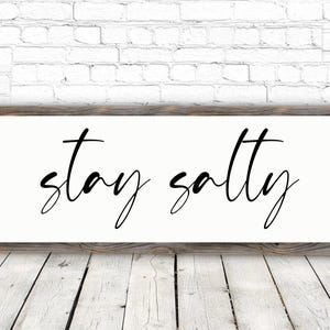 May include: A rectangular wooden sign with the words "stay salty" in black cursive script. The sign has a white background and is framed by a rustic, weathered wood. The sign is mounted on a white brick wall.