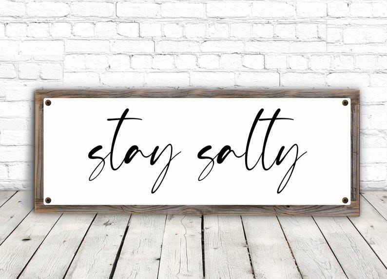 May include: A rectangular wooden sign with the words "stay salty" in black cursive script. The sign has a white background and is framed by a rustic, weathered wood. The sign is mounted on a white brick wall.