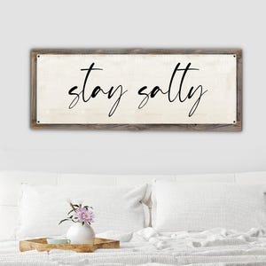 May include: A rectangular wooden sign with the words "stay salty" in black cursive lettering. The sign has a light beige background and a rustic, weathered wood frame. The sign is mounted above a bed with white pillows and a decorative tray.
