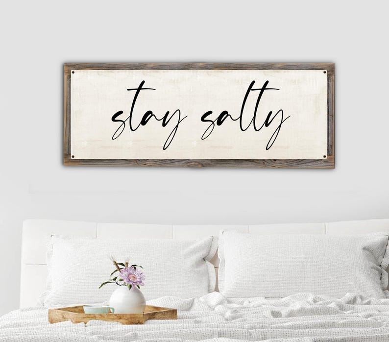May include: A rectangular wooden sign with the words "stay salty" in black cursive lettering. The sign has a light beige background and a rustic, weathered wood frame. The sign is mounted above a bed with white pillows and a decorative tray.