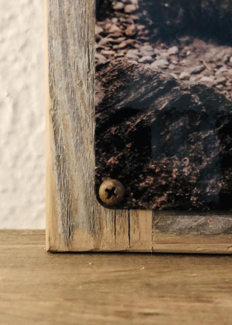 May include: Close-up of a framed photograph with a rustic, weathered wooden frame. The frame is a light brown color with visible wood grain and a brass screw. The photograph depicts a rocky landscape.
