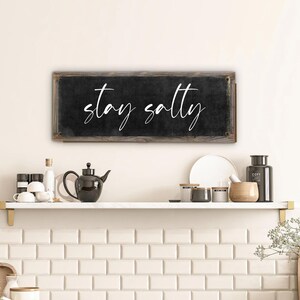 May include: A rectangular black sign with the words "stay salty" in white script. The sign has a rustic, wooden frame and is mounted on a white wall above a shelf with kitchen items.