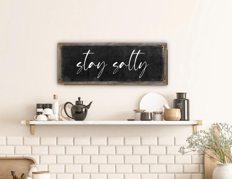 May include: A rectangular black sign with the words "stay salty" in white script. The sign has a rustic, wooden frame and is mounted on a white wall above a shelf with kitchen items.