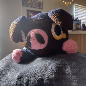 May include: A crocheted plush toy pig with brown, pink, blue, white, and yellow yarn. The pig is lying on a gray surface.