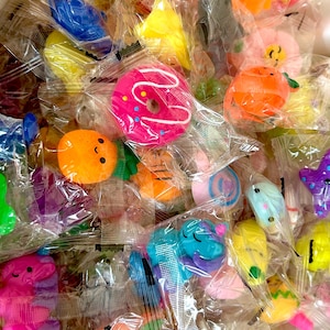 May include: A collection of colorful, individually wrapped squishy toys. The assortment includes a pink donut, orange fruits, blue animals, and other shapes. Each toy is sealed in clear plastic.