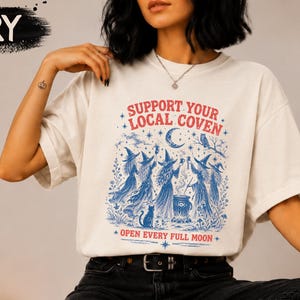 May include: An ivory short-sleeve t-shirt featuring a blue and red graphic print. The text reads "SUPPORT YOUR LOCAL COVEN" and "OPEN EVERY FULL MOON" surrounding an illustration of five witches gathered around a cauldron with a cat and moon.