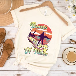 Op de afbeelding: Een wit T-shirt met een afbeelding van twee vrouwen op een boot met de tekst "Boats 'N Hoes" in een retro-lettertype.