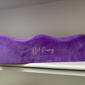 Op de afbeelding: Paarse beenkussen met een wit geborduurd logo met de tekst "RM Recovery WASHINGTON DC".