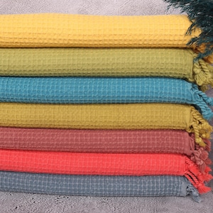 Puede incluir: Una pila de siete toallas de tejido de gofre en varios colores, incluyendo amarillo, verde pistacho, azul petróleo, mostaza, ladrillo oscuro, rojo y gris oscuro. Cada toalla tiene una franja en el extremo.