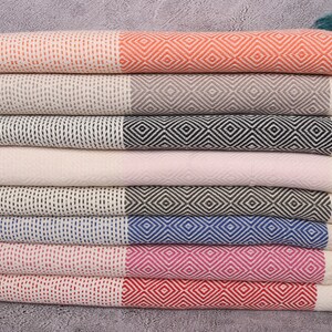 May include: A stack of eight Turkish towels in various colours, including orange, light brown, black, light pink, brown, sea blue, fuchsia, and red. Each towel has a white background with a diamond pattern in the colour of the towel.