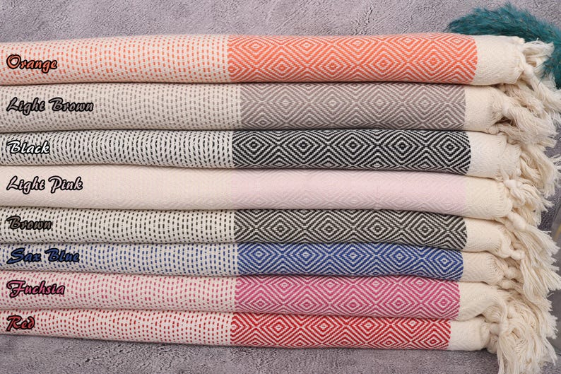 May include: A stack of eight Turkish towels in various colours, including orange, light brown, black, light pink, brown, sea blue, fuchsia, and red. Each towel has a white background with a diamond pattern in the colour of the towel.