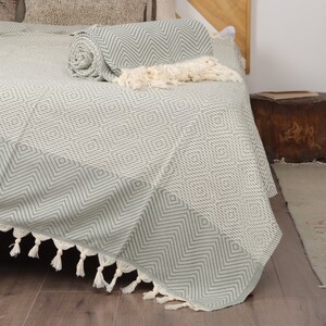 May include: A light green and white patterned bedspread with a geometric design. The bedspread is folded over a bed, with a rolled-up section and a decorative fringe. The bedspread is made of a soft, woven material.