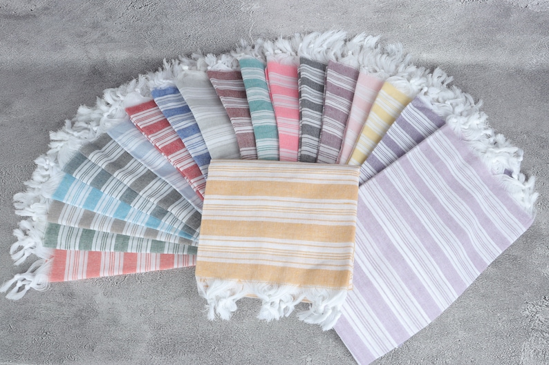May include: A collection of 15 striped Turkish towels in various colors, including red, blue, green, yellow, pink, and purple. The towels are arranged in a fan shape and have white fringe.