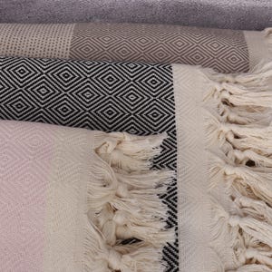 May include: A stack of three Turkish towels with fringe. The top towel is light brown with a diamond pattern, the middle towel is black and white with a diamond pattern, and the bottom towel is light pink with a diamond pattern.
