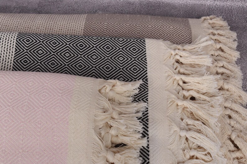 May include: A stack of three Turkish towels with fringe. The top towel is light brown with a diamond pattern, the middle towel is black and white with a diamond pattern, and the bottom towel is light pink with a diamond pattern.
