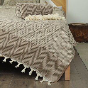 May include: A beige and brown geometric patterned bedspread with a rolled-up blanket and decorative tassels. The bedspread features a diamond and zigzag design. The bed frame is made of light-colored wood.