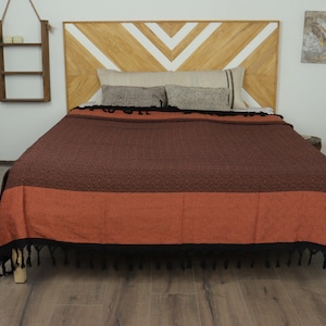 May include: A brown and orange patterned blanket with black fringe is draped over a bed. The blanket has a textured design and is made of a soft material. The headboard is made of wood with a chevron pattern.