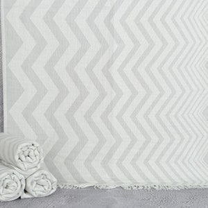 Puede incluir: Toalla de playa con estampado de chevrón gris claro y blanco con borde con flecos. Tres toallas enrolladas con el mismo patrón están apiladas frente a la toalla desplegada. Las toallas están hechas de un material suave y ligero.