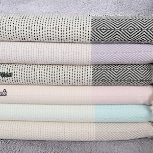 Puede incluir: Una pila de seis toallas de algod&oacute;n tejido en varios colores pastel. Cada toalla tiene un borde con estampado geom&eacute;trico y una borla. Los colores incluyen negro, lila, verde caqui, rosa claro, menta y beige.