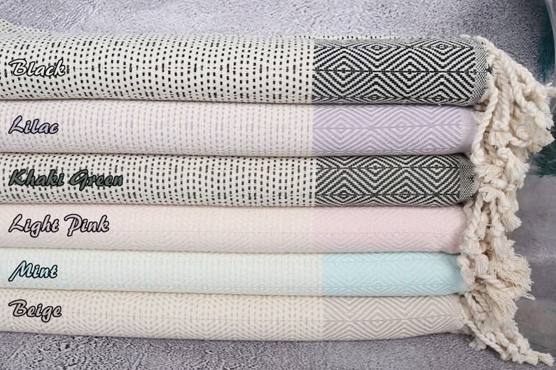 Puede incluir: Una pila de seis toallas de algod&oacute;n tejido en varios colores pastel. Cada toalla tiene un borde con estampado geom&eacute;trico y una borla. Los colores incluyen negro, lila, verde caqui, rosa claro, menta y beige.