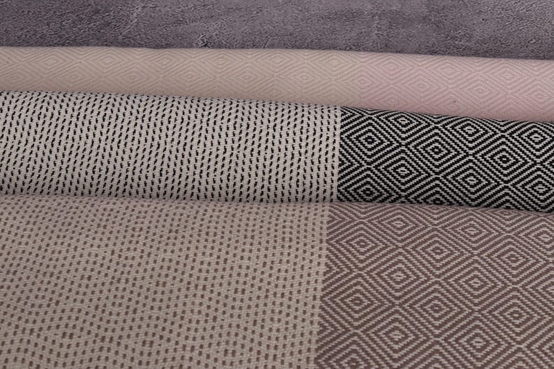 May include: A stack of four woven towels with different patterns. The top towel is pink with a diamond pattern, the second towel is white with a black diamond pattern, the third towel is white with a black wavy line pattern, and the bottom towel is beige with a brown diamond pattern.