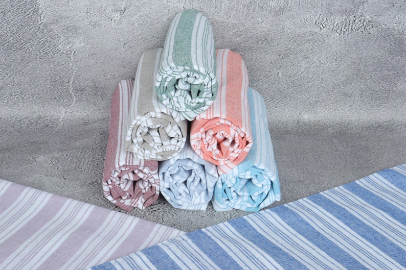 May include: A stack of rolled striped towels in various colors, including pink, blue, green, and beige. The towels are arranged in a pyramid shape on a gray textured surface.