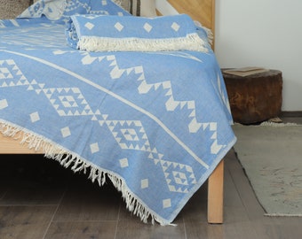 May include: A light blue and white patterned blanket with a geometric design, fringed edges, and a rolled-up matching blanket on a bed. The blanket is draped over a wooden bed frame.