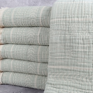 May include: A stack of light green cotton towels with white stripes and fringed edges. The towels have a textured, crinkled appearance and a subtle geometric pattern. The towels are folded neatly, showcasing their soft, inviting texture.