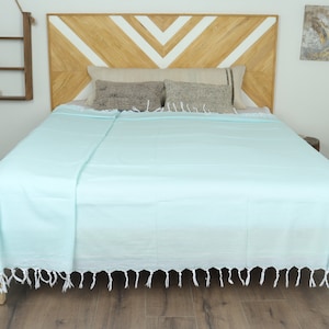 May include: A light blue bedspread with white fringe is draped over a bed. The bed has a wooden headboard with a chevron design. Decorative pillows rest on the bed. A small wooden shelf and a potted plant are visible.