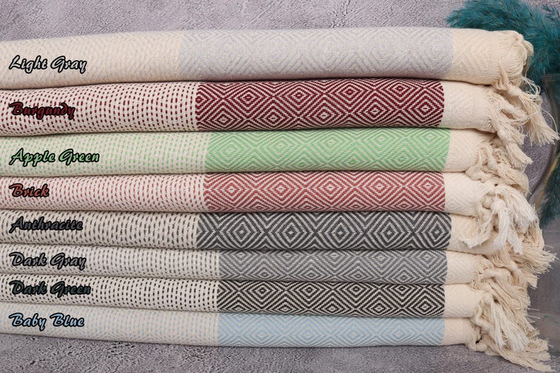 May include: A stack of eight Turkish towels in various colours, including light grey, burgundy, apple green, brick, anthracite, dark grey, dark green, and baby blue. Each towel has a diamond pattern and a fringe.