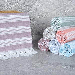 May include: A stack of six rolled striped towels in various colors, including pink, blue, green, and gray. The towels have white fringe and are arranged in a pyramid shape. A single towel is lying flat in a wicker basket.