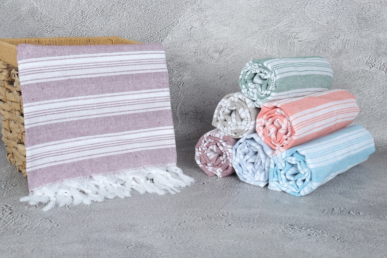 May include: A stack of six rolled striped towels in various colors, including pink, blue, green, and gray. The towels have white fringe and are arranged in a pyramid shape. A single towel is lying flat in a wicker basket.