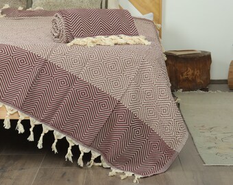 May include: A burgundy and beige geometric patterned throw blanket with tassels. The blanket is folded and rolled on a bed. The design features diamond shapes and zigzags. The tassels are cream-colored.