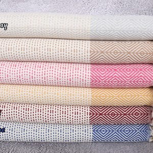 Puede incluir: Una pila de seis toallas de algod&oacute;n tejido en varios colores. Las toallas est&aacute;n etiquetadas como "Light Gray", "Brown", "Fuchsia", "Mustard", "Burgundy" y "Sax Blue". Cada toalla tiene un patr&oacute;n de diamantes en un extremo y flecos.