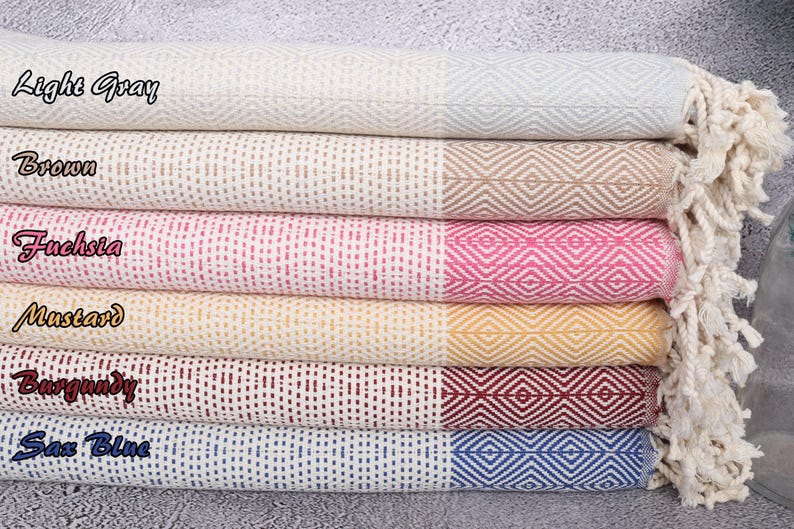 Puede incluir: Una pila de seis toallas de algod&oacute;n tejido en varios colores. Las toallas est&aacute;n etiquetadas como "Light Gray", "Brown", "Fuchsia", "Mustard", "Burgundy" y "Sax Blue". Cada toalla tiene un patr&oacute;n de diamantes en un extremo y flecos.