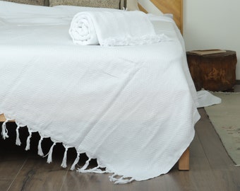 May include: A white herringbone-patterned blanket with a fringe detail is draped over a bed. A rolled-up blanket and a folded blanket are also on the bed. The bed frame is made of light-colored wood.