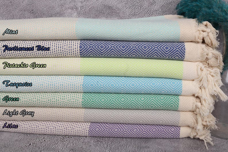 May include: A stack of seven Turkish towels in various colours, including mint, turquoise, pistachio green, light grey, and lilac. Each towel has a white border and a diamond pattern.