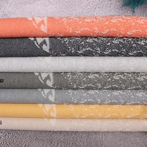 May include: A stack of seven towels in various colors, including orange, navy, light gray, khaki green, dark gray, mustard, and light beige. The towels have a white geometric pattern and fringe trim.