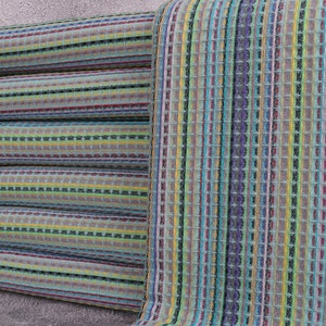 May include: A stack of colorful striped towels with a waffle weave texture. The towels feature a mix of vertical stripes in shades of blue, green, yellow, and purple. The edges have knotted fringe.