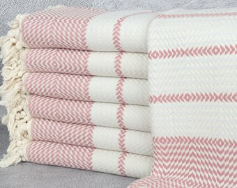 May include: A stack of white and pink patterned towels with fringed edges. The towels feature a geometric design in pink and white, with a textured weave. The towels are neatly folded and stacked, showcasing the soft texture.