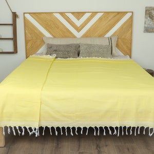 May include: A bright yellow bedspread with white fringe is draped over a bed. The bed has a wooden headboard with a chevron design. Decorative pillows and a neutral-toned blanket are on the bed. The room has a neutral color palette.