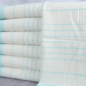 May include: A stack of six white waffle-weave towels with light blue stripes and trim. The towels have a fringed edge and are neatly folded, showcasing a clean and minimalist aesthetic. Ideal for bathroom decor.