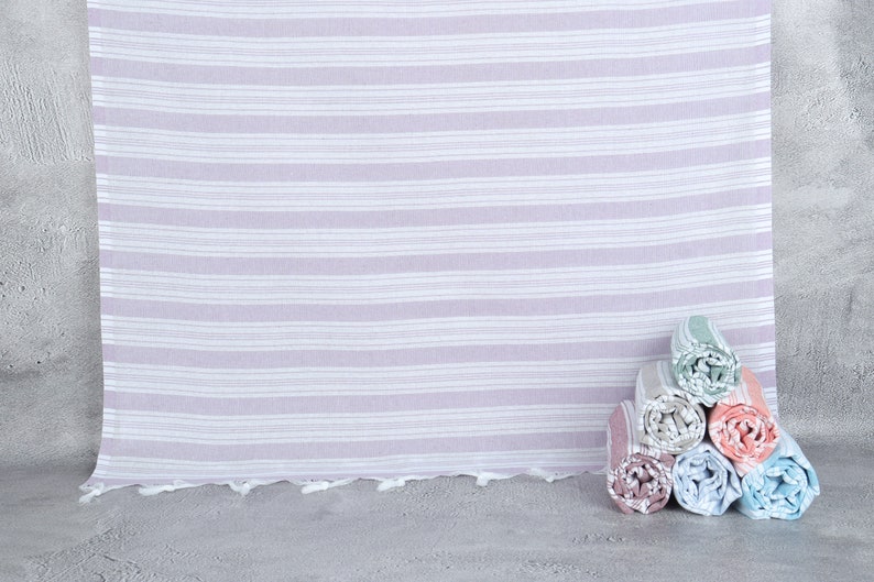 May include: A stack of six rolled towels in various colors, including pink, blue, green, and peach, with a white and light purple striped towel in the background.