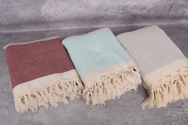 May include: Three Turkish towels with fringe. One towel is red with a diamond pattern, one is light blue, and one is grey.
